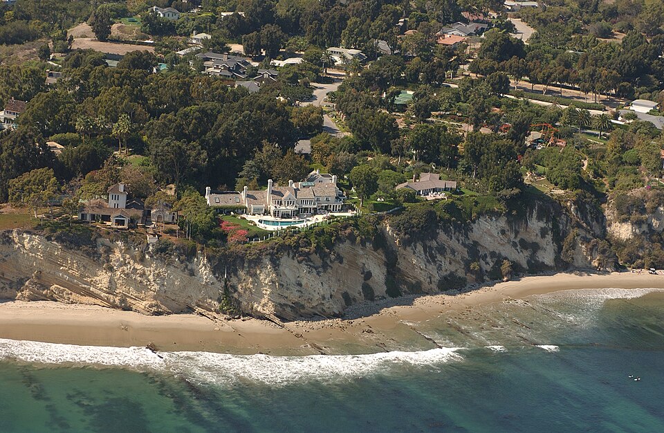 Aerial view of a large mansion estate on a Malibu cliff, with the Pacific Ocean below and dense greenery inland.