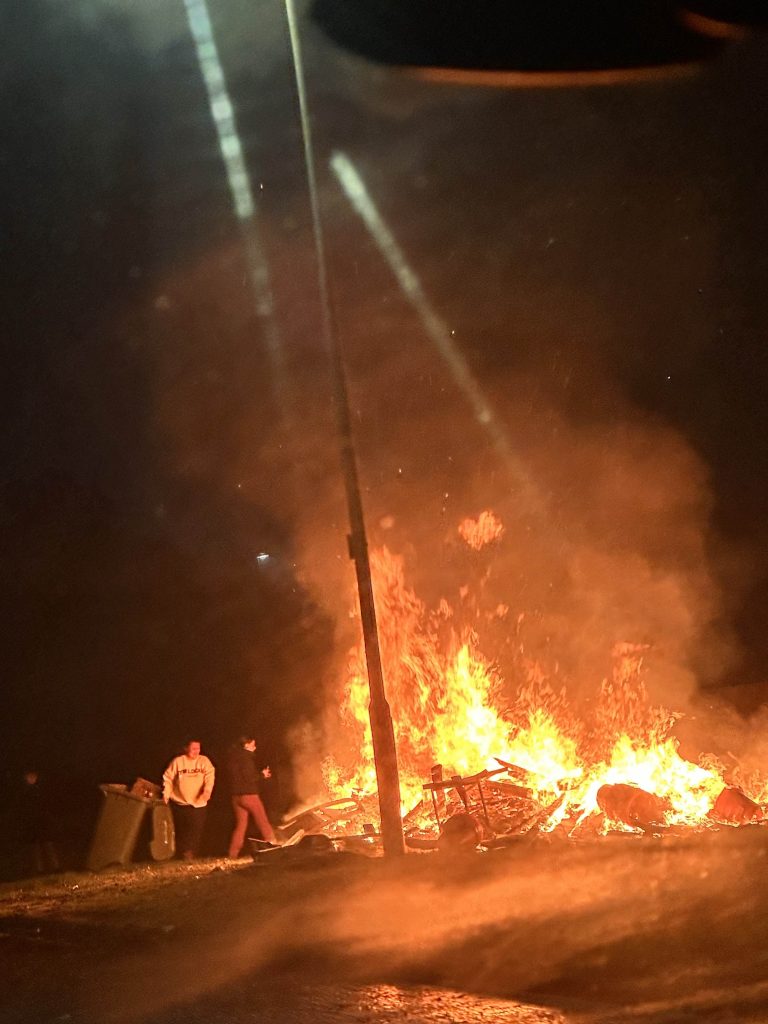 Large community bonfire at night on a council estate, residents standing nearby as flames and smoke light the street