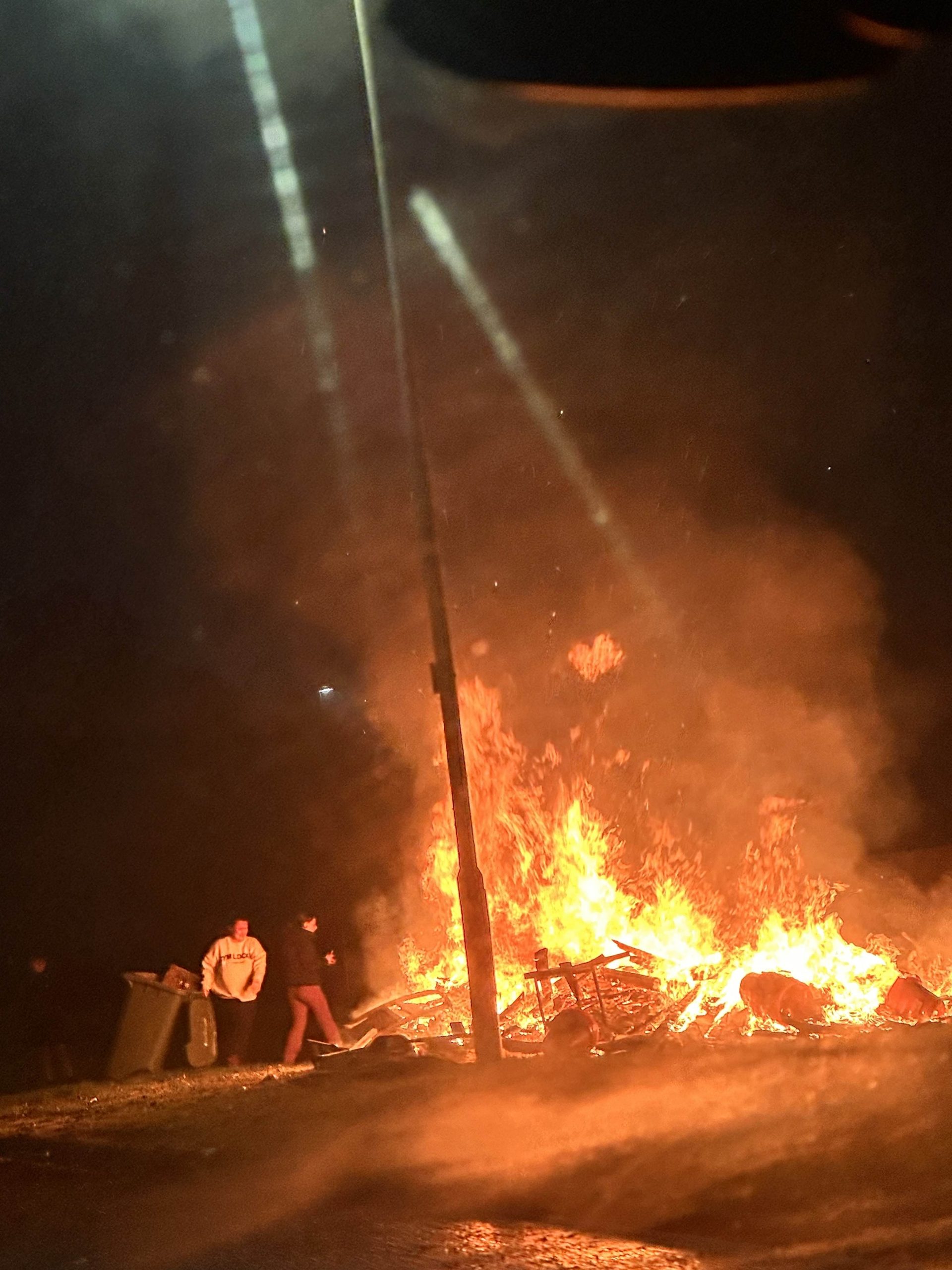 Large community bonfire at night on a council estate, residents standing nearby as flames and smoke light the street