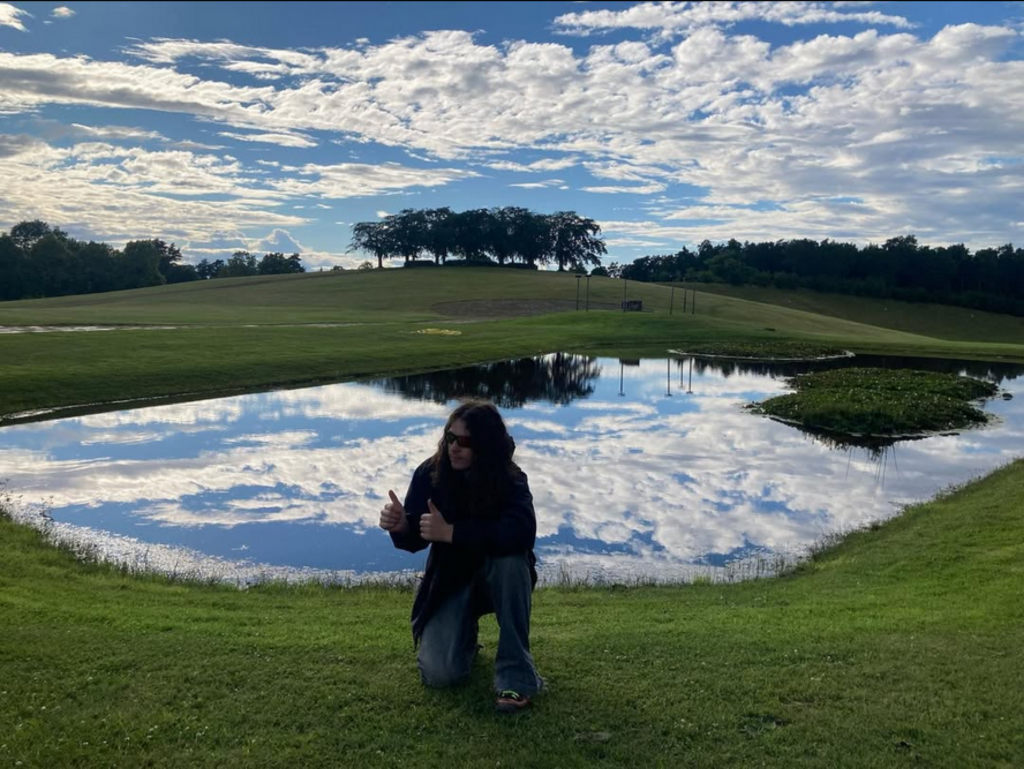 Bladee kneeling beside a reflective pond in a grassy landscape, giving a thumbs up