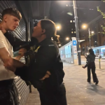 Female police officer restraining a young man against a closed shop shutter on Broad Street in Birmingham at night.