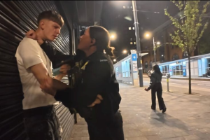 Female police officer restraining a young man against a closed shop shutter on Broad Street in Birmingham at night.