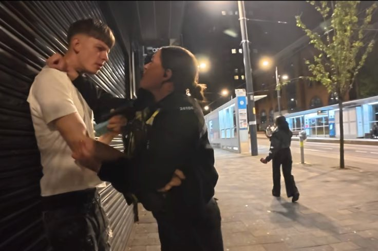 Female police officer restraining a young man against a closed shop shutter on Broad Street in Birmingham at night.